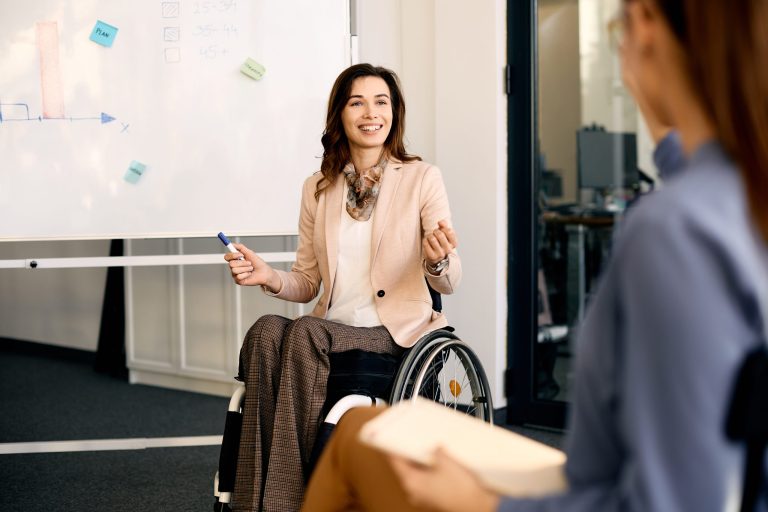 A businesswoman with long brown hair and a friendly smile, dressed in a light pink business suit and using a wheelchair, leads a meeting with a whiteboard behind her, conveying expertise and leadership.