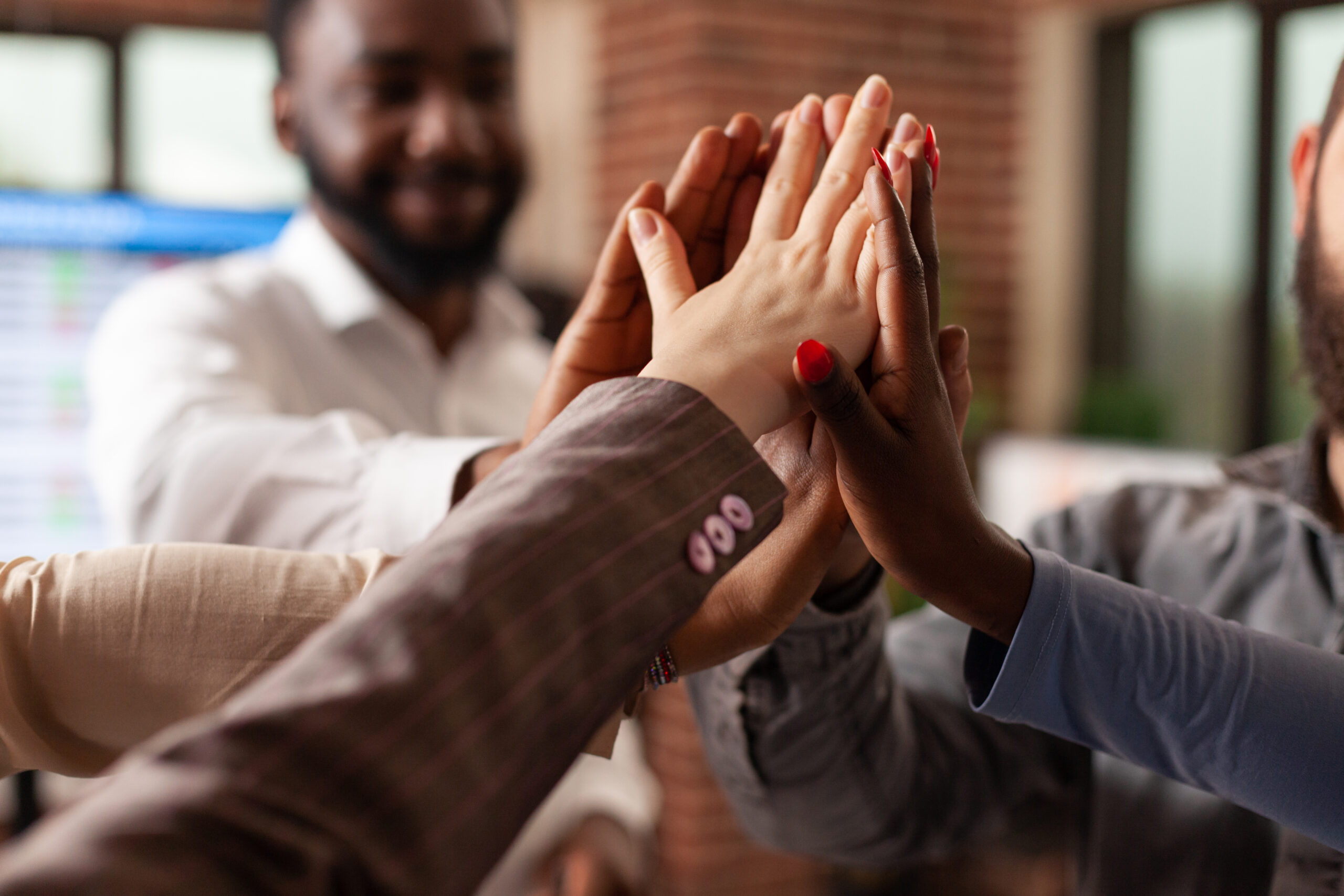 In a startup office, a diverse, smiling business team celebrates a successful partnership with a giant high five, expressing excitement and achievement in business collaboration.